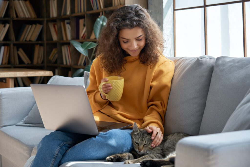 Happy hispanic teen girl relaxing on couch holding laptop playing with cat.