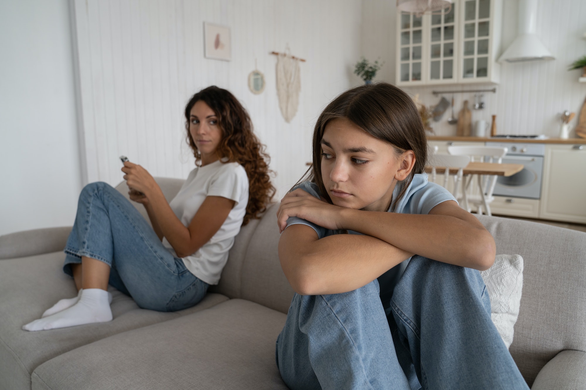 Offended teen girl feeling sad after argument with parent, sitting separately with mother on sofa