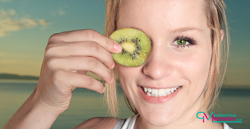 Teenage girl holding a slice of kiwi fruit to her eye.